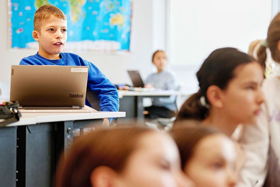 Arbian, ein Junge, sitzt an seinem Pult mit aufgeklapptem Laptop. Auf dem Foto sind weitere Mitschülerinnen und -schüler zu sehen. 