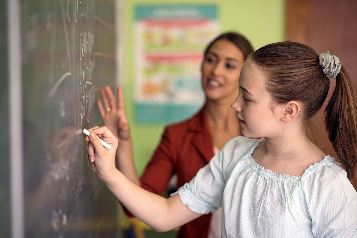 Ein Mädchen schreibt an die Wandtafel. Hinter ihr steht die Lehrerin.