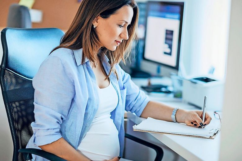 1199485417 Am Bürotisch sitzt eine schwangere Frau, die sich vor dem Computer Notizen macht.