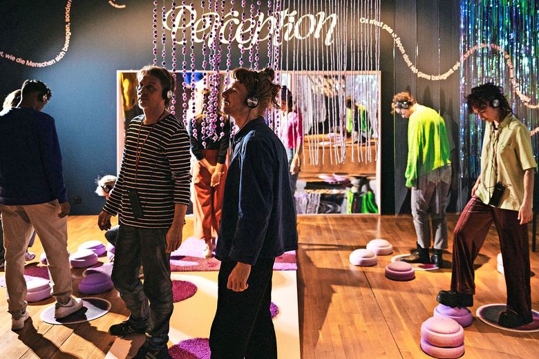 Mehrere Menschen stehen in einem Raum. Sie tragen Kopfhöher. Von der Decke hängt ein Schriftzug herunter, der "Perception" buchstabiert.