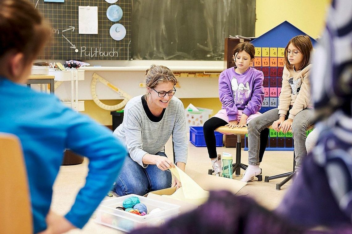 Kinder sitzen auf Stühlen in einem Kreis. In der Mitte des Kreises kniet eine Frau auf dem Boden. Vor ihr liegen Stoffe und Wollknäuel.