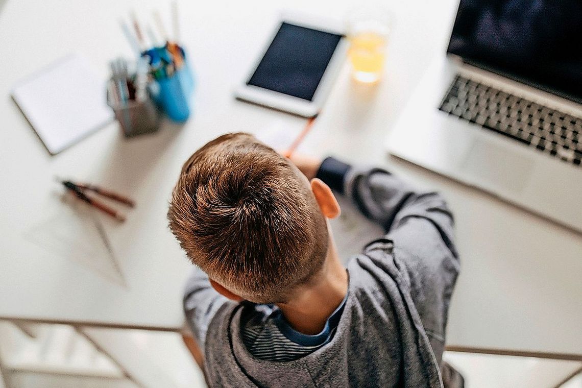 Junge sitzt an seinem Bürotisch, auf dem es sowohl Stift und Papier wie ein Computer und ein Tablet hat.