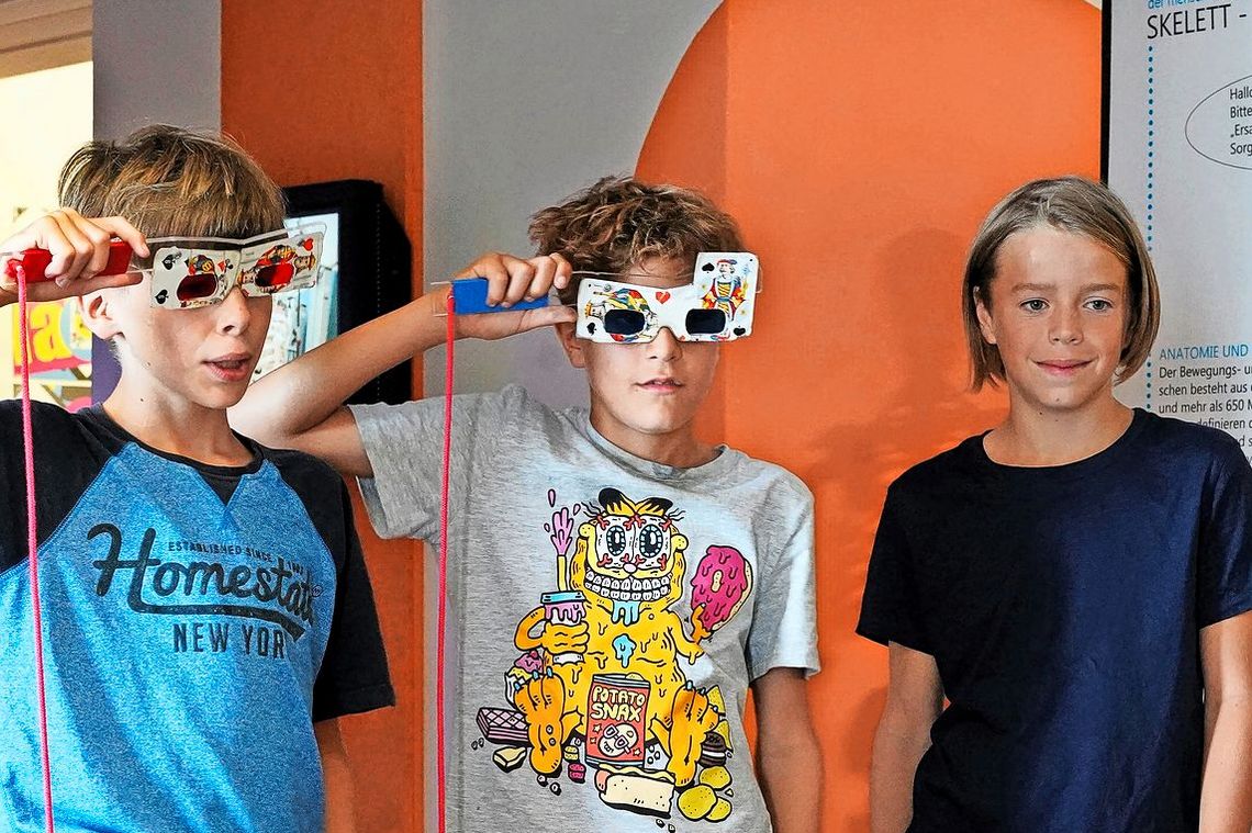 Drei Jungen stehen vor einer Wand. Zwei von ihnen haben je eine Brille mit einem Griff daran aufgesetzt.