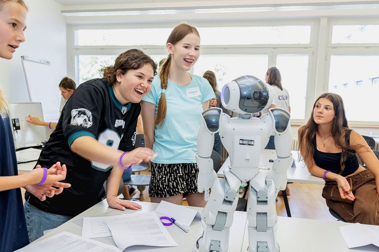 Mehrere Mädchen stehen um einen Tisch und betrachten lachend einen Roboter, der dort steht.