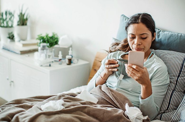 Frau liegt einer Tasse und einer Erkältung im Bett und schaut auf ihr Smartphone.
