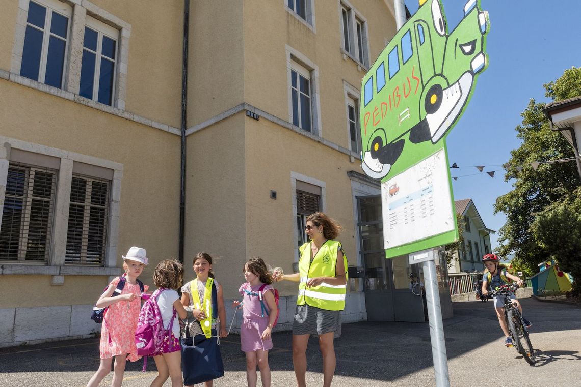 Kinder und eine Mutter beim Schild der Pedibus-Haltestelle vor dem Schulhaus