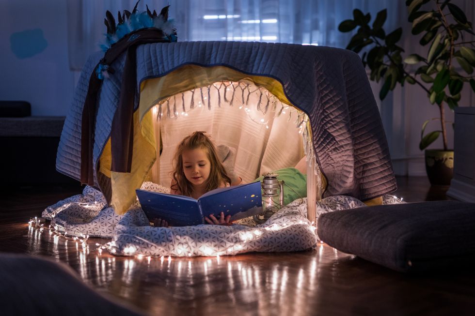 Cute girl reading children's book in a tent at home. Mädchen liest in einem mit Lichterketten dekorierten Zelt