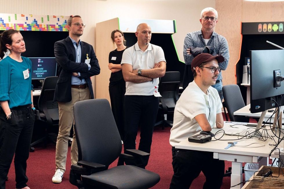 Patrick Schilling steht am Computer umgeben von Arbeitskolleginnen und -kollegen.