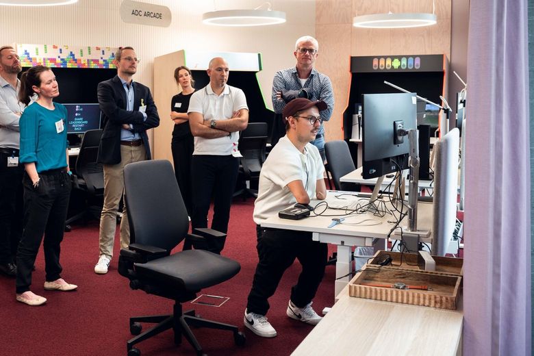 Patrick Schilling steht am Computer umgeben von Arbeitskolleginnen und -kollegen.