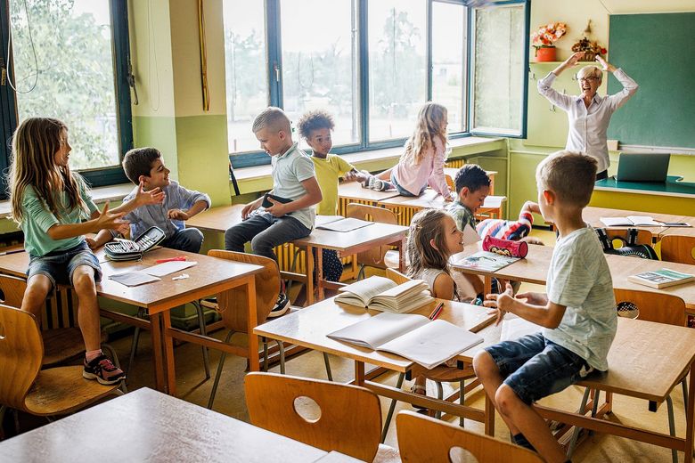 1150473625 Schulkinder, die auf Pulten sitzen und schwatzen. Im Hintergrund eine überfordert die Hände über dem Kopf zusammenschlagende Lehrerin.