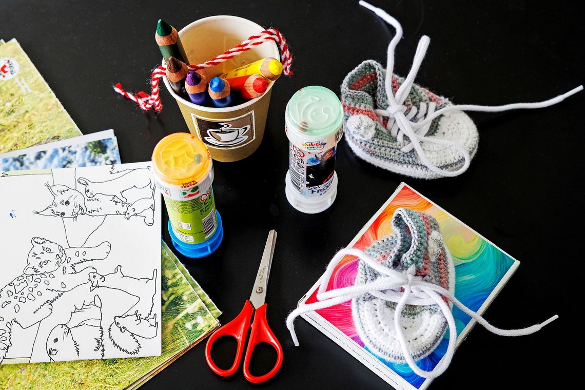 Verschiedene Gegenstände liegen auf einer Oberfläche. Bei den Gegenständen handelt es sich um eine Schere, Papier mit ausmalbaren Zeichnungen, Stiften, Seifenblasen-Behältern und zwei Schuhen in Kleinkindgrösse.