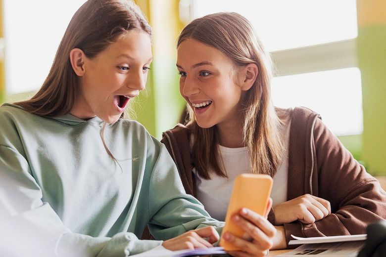 Eine Jugendliche betrachtet mit offenem Mund etwas auf einem Smartphone. Eine weitere Jugendliche beobachtet sie grinsend.
