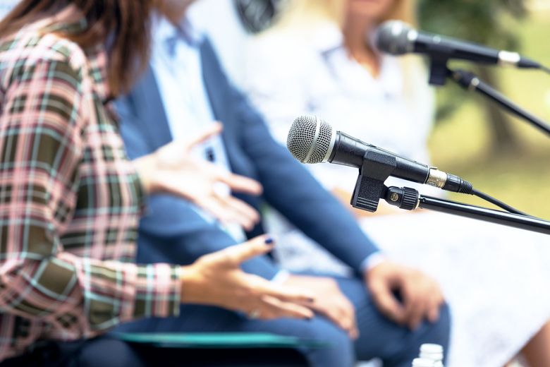Ein Mikrofon und gestikulierende Hände im Hintergrund deuten eine Diskussion an.