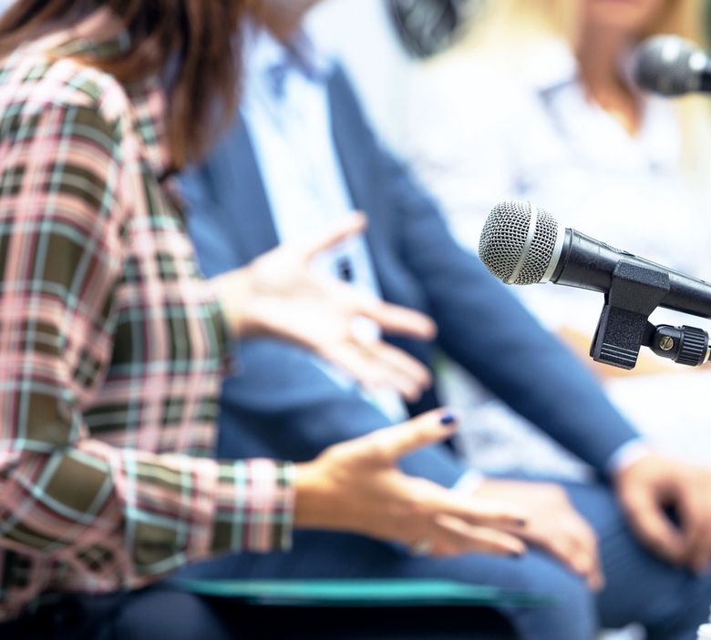 Ein Mikrofon und gestikulierende Hände im Hintergrund deuten eine Diskussion an.