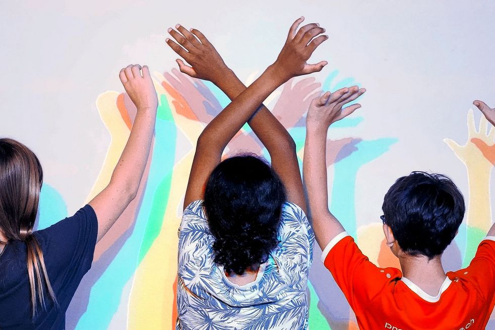 Drei Kinder stehen an einer Wand und haben die Arme erhoben, während sie ihre bunten Schatten an der Wand betrachten.