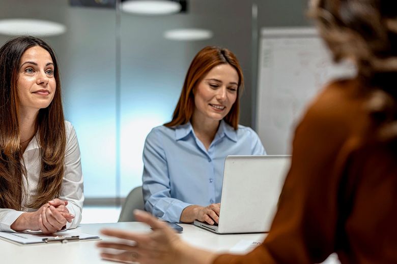 Zwei Frauen sitzen einer dritten Person am Tisch gegenüber.