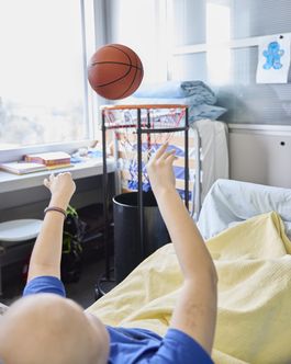 Ein Junge in einem Spitalbett wirft einen Basketball in ein Netz.