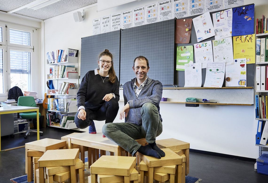 Anna Bühler und Peter Liechti sitzen im Schulzimmer auf ein paar Kindertischen.