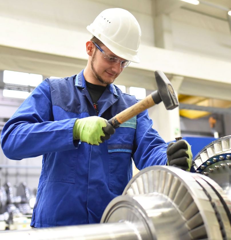 Junger Mann trägt einen Helm und arbeitet mit einem Hammer an einer Maschine.