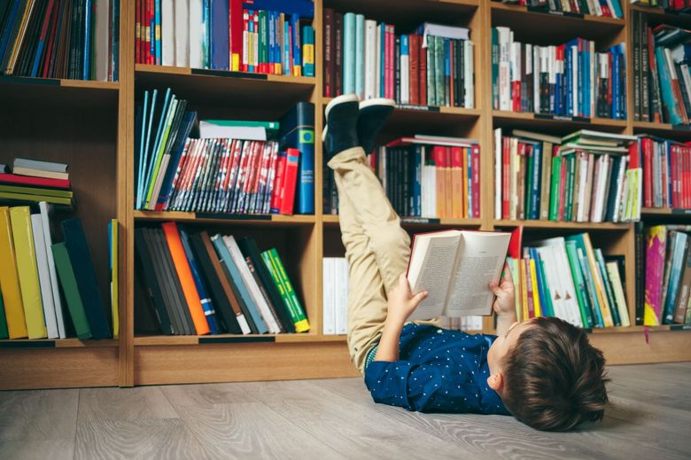 In einer Bibliothek liegt ein Kind lesend auf dem Rücken und lehnt seine Beine ans Bücherregal.