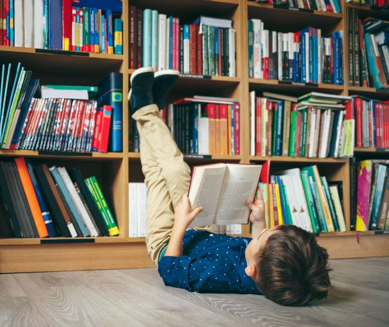 In einer Bibliothek liegt ein Kind lesend auf dem Rücken und lehnt seine Beine ans Bücherregal.