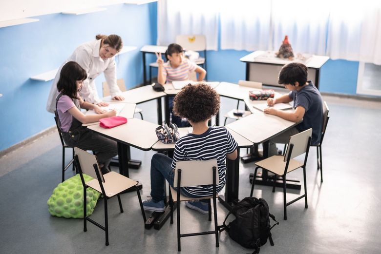 Schulkinder sitzen an rundem Tisch und werden von der Lehrerin betreut.