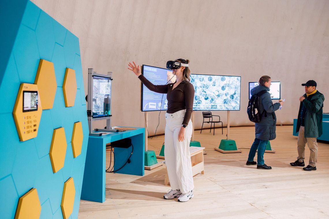 Eine junge Frau steht vor einem Bildschirm und trägt eine VR-Brille.
