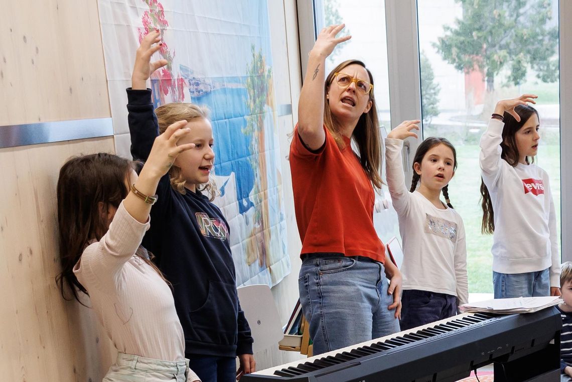 Reportage Tagesschule Prêles Florine Némitz am E-Piano umgeben von Kindern. Zusammen üben sie die Gebärdensprache.