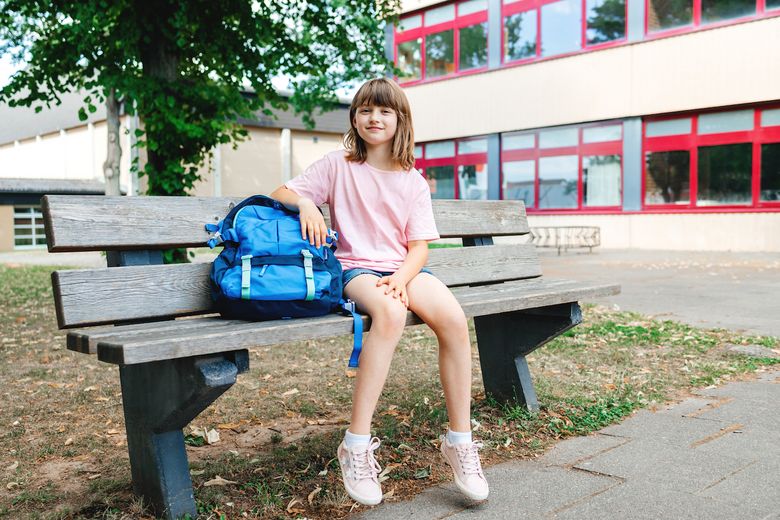 Back to school. A schoolgirl teenager sits on a bench next to a schoolbag in front of the school, smiling and looking at the camera.