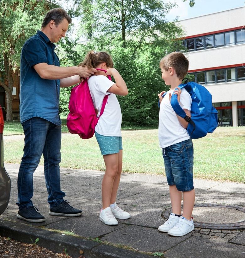 Auto vor einem Schulhaus Vater hilft Tochter beim Anziehen eines Schulrucksacks Sohn schaut zu