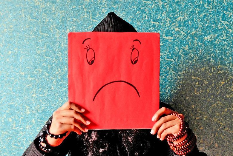 eine jugendliche Person hält ein Blatt vor ihr Gesicht. Auf das Blatt ist ein Gesicht mit hängendem Mund gezeichnet.