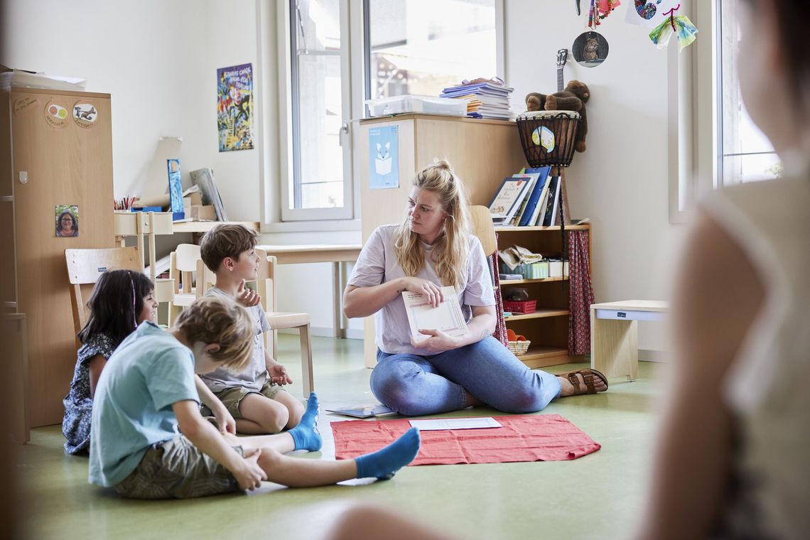 Eine Lehrerin und Kinder sitzen am Boden eines Klassenzimmers.