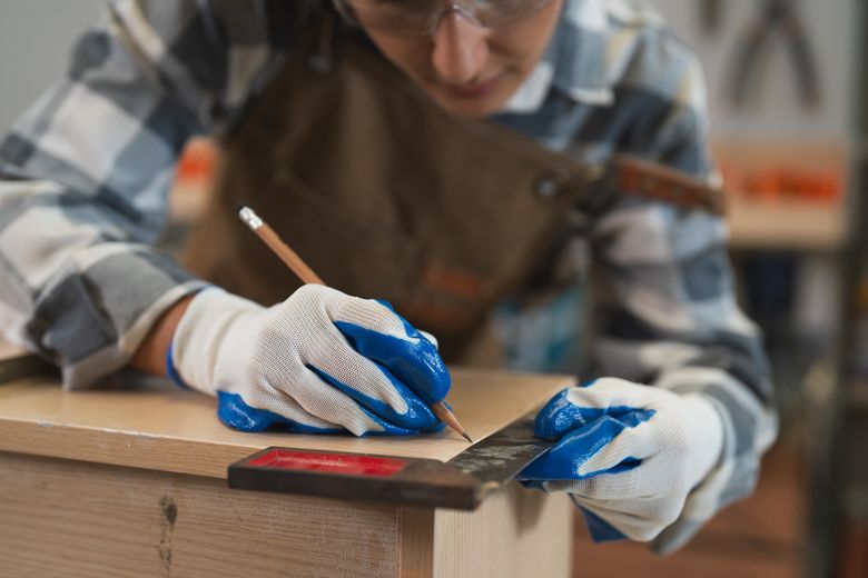 Female carpenter hands wear gloves using square ruler tool measuring length of wooden plank in carpentry workshop. Person vermisst ein Stück Holz.
