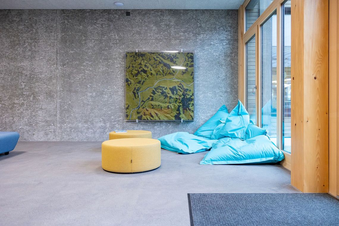 Ein Raum aus Beton mit verschiedenen Sitzgelegenheiten und einem raumhohen Fenster.