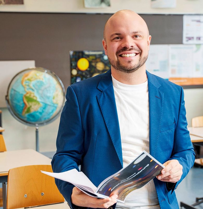 Porträt des Lehrers Thomas Klaper, der ein Buch in den Händen hält und in einem Klassenzimmer steht.