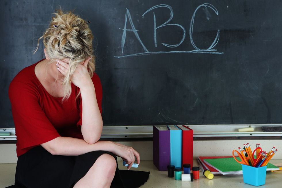 Frustrated elementary teacher Eine Person sitzt vor einer Wandtafel und hat die Stirn in eine Hand gelegt.