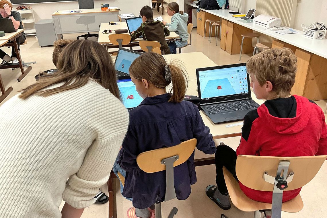 Eine Person mit langen Haaren beugt sich zu einem Laptop, an dem ein Kind sitzt. Daneben sitzt ein weiteres Kind am Laptop.