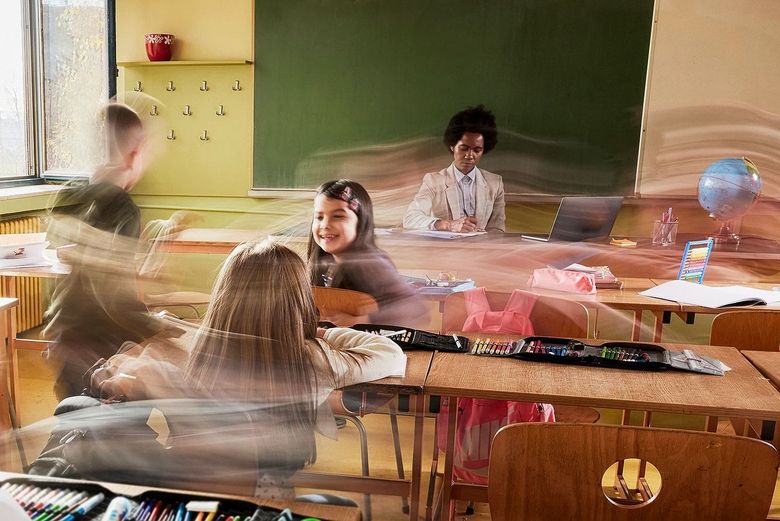 In einem Klassenzimmer sitzt eine Frau an einem Schreibtisch vor einer Wandtafel. Vor dem Schreibtisch stehen Schulbänke, zwischen denen drei Kinder stehen. Ihre Bewegungen sind verzerrt dargestellt, was den Eindruck von viel Aktivität und Geschwindigkeit vermittelt.