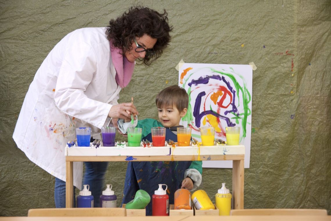 Betreuerin hilft einem Kind, das vor sich Gläser mit flüssiger Farbe hat, zu malen.