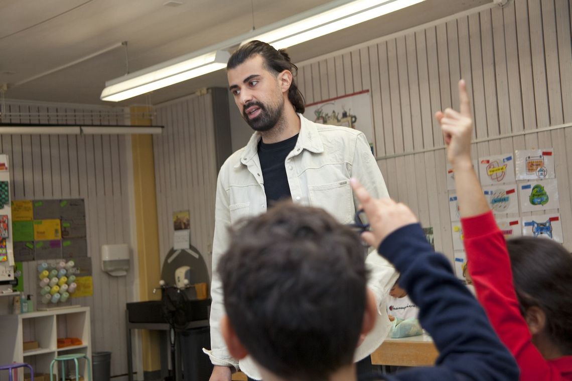 Primarlehrer Özcan Azak beim Unterrichten. Er ist umringt von Schülerinnen und Schülern. Einige strecken den Finger auf.