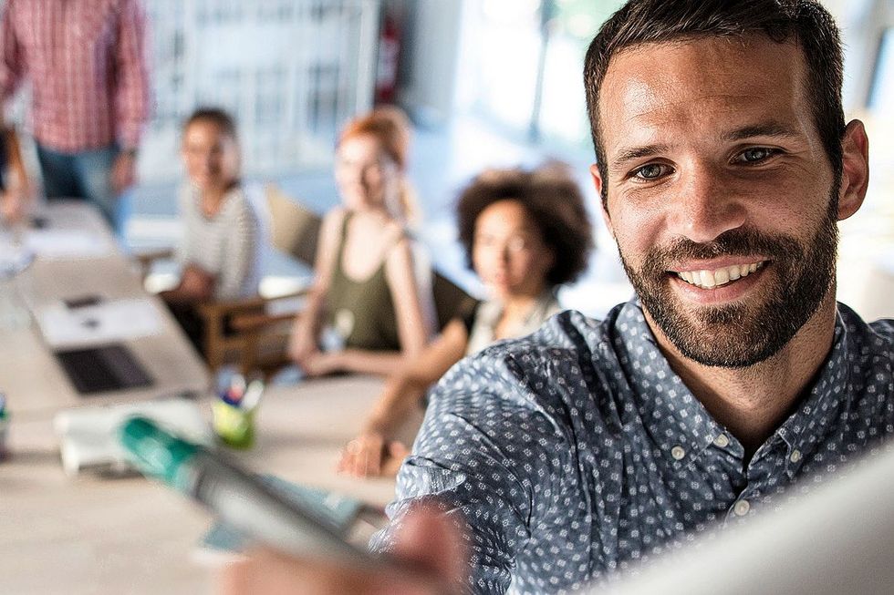 Ein lächelnder Mann schreibt etwas auf eine Leinwand. Hinter ihm sitzen mehrere Personen an einem Tisch.