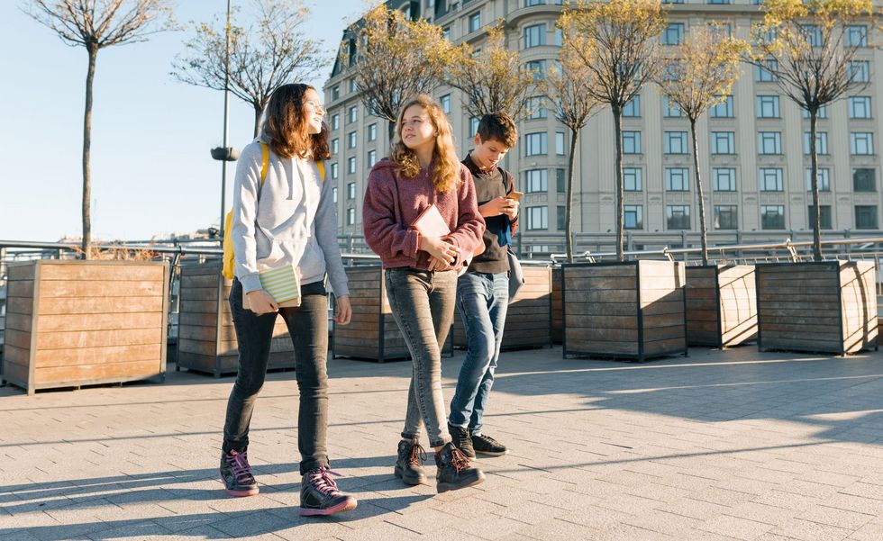 Outdoor portrait of teenage students with backpacks walking and talking. City background, golden hour Drei Jugendliche schlendern über einen Platz.