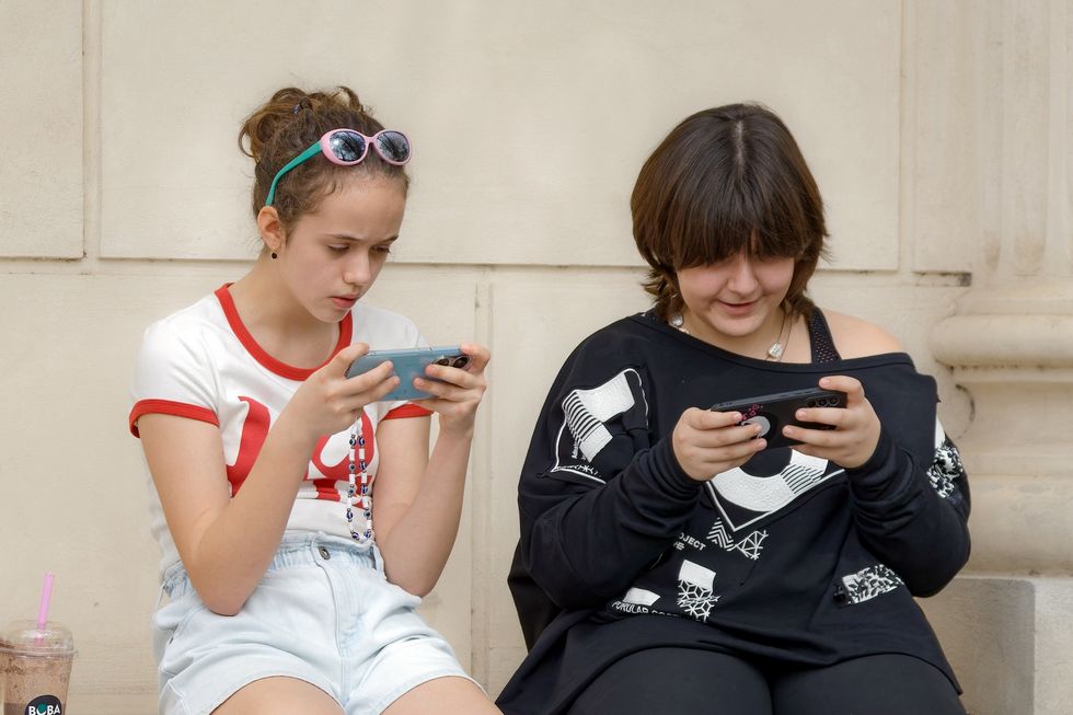 Zweimädchen sitzen auf einer Mauer und starren auf ihre Handys.