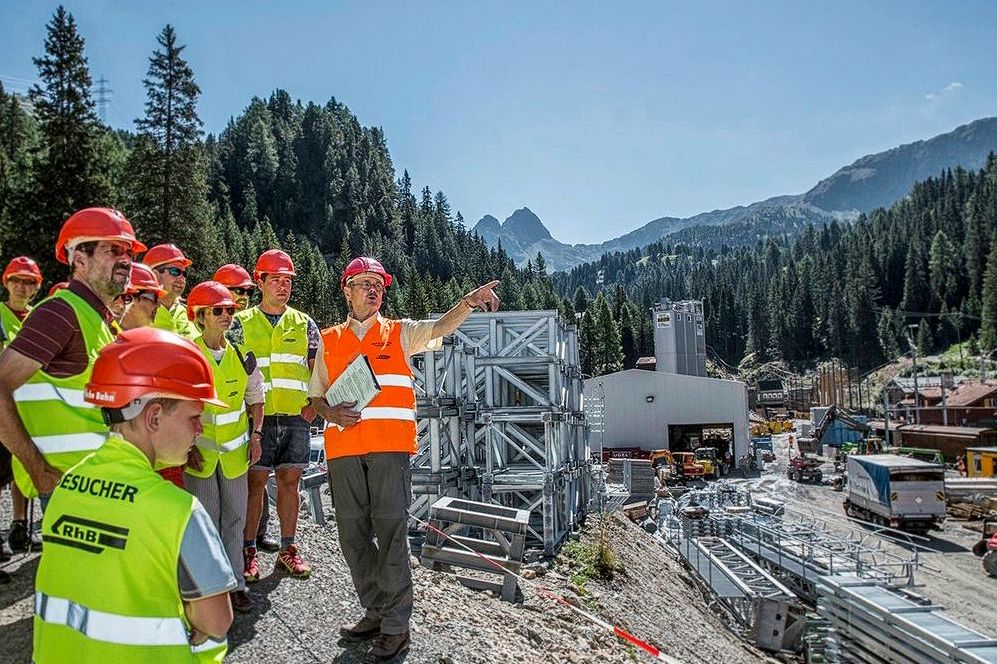 Personen in Signalwesten und Bauhelmen besuchen eine Baustelle.