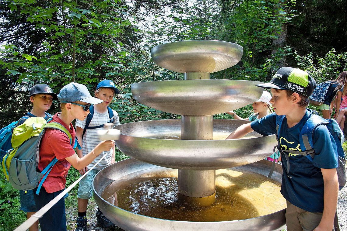 Ein paar Kinder bewundern einen Brunnen aus Metall, an dem ein Schlaghammer hängt.