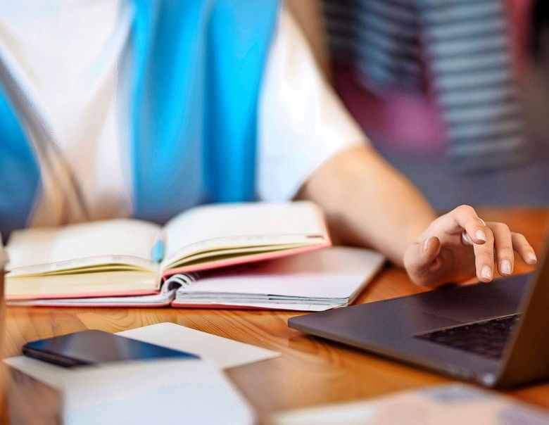 Eine Person hat zwei offene Bücher vor sich auf dem Tisch liegen und bedient einen Laptop.