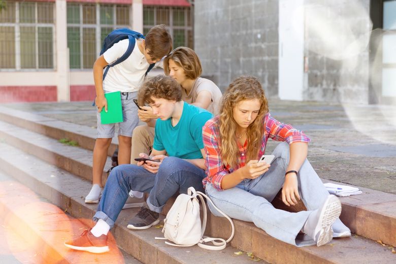 Drei Jugendliche sitzen vor einem Gebäude auf einer Treppe. Ein vierter Jugendlicher steht dabei. Alle schauen auf Smartphones.